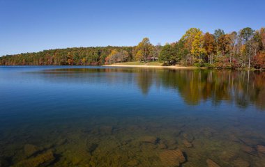 Indian Boundary Lake in autumn, vibrant red, yellow, and green trees reflect in the calm blue water and a lakeside swimming area visible. Accessed from the Cherohala Skyway in Tennessee. Horizontal clipart