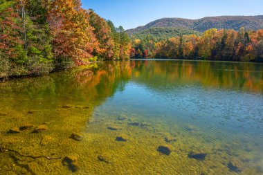 The Indian Boundary Lake in autumn, with vibrant red, yellow, orange, and green trees along the shoreline and reflected in the water. The Unicoi mountains are in the background. Horizontal clipart