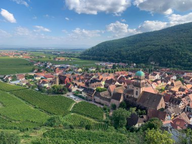 Kaysersberg, Alsace, Fransa 'nın havadan görünüşü. Üzüm bağları ve parlak gökyüzünün altındaki yeşil tepeler. Köyde geleneksel evler ve doğa ile çevrili tarihi bir kilise yer alıyor.