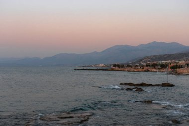 Hersonissos, Girit kıyı şeridi. Sahildeki sahil kenti ve dağları görüyor. Gün batımında okyanus üzerinde.