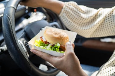 Asyalı kadın sürücü arabada hamburger tutuyor ve yiyor..