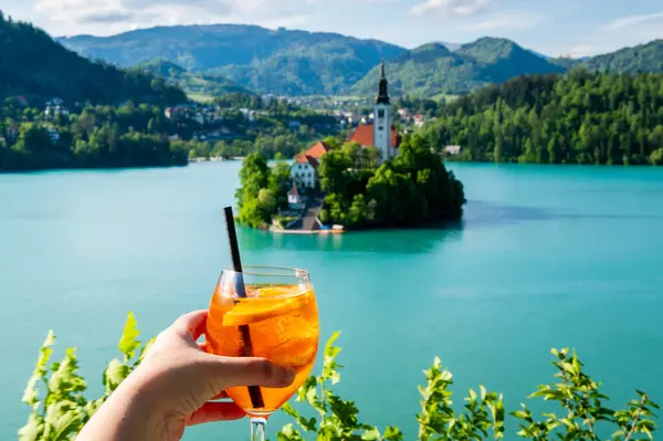 Slovenya, Bled Gölü 'nün önünde elinde turuncu Spritz kokteyli olan bir kadın kadeh kaldırıyor.