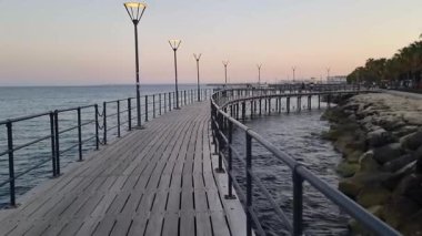 Kıbrıs Rum Kesimi 'nin Limassols Molos kentinde gün batımında Akdeniz kıyısı boyunca uzanan sakin bir ahşap iskele. Fenerlerle aydınlatılan huzurlu akşam sahnesi kıyı şeridini vurguluyor.