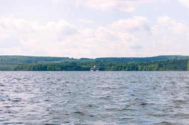 Büyük bir tekne sakin bir gölde yelken açıyor. Gökyüzü bulutlu ve su durgun.