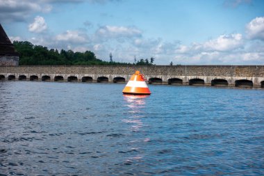 Büyük bir su kütlesinin yanında bir şamandıra yüzüyor. Şamandıra turuncu ve beyaz.