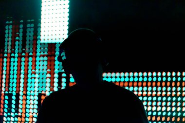 Işıklardan oluşan bir duvarın önünde bir adam duruyor. Işıklar farklı renk ve şekillerde, dinamik ve görsel olarak ilginç bir sahne oluşturuyor. Adam kulaklık takıyor.