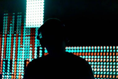 Işıklardan oluşan bir duvarın önünde bir adam duruyor. Işıklar farklı renk ve şekillerde, dinamik ve görsel olarak ilginç bir sahne oluşturuyor. Adam kulaklık takıyor.