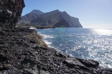 Arka planda büyük bir su kütlesi olan kayalık bir kıyı şeridi. Su sakin ve gökyüzü berrak.