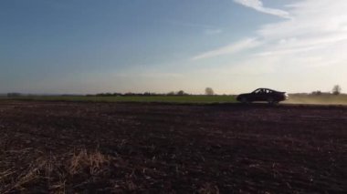 Siyah bir araba tarlada toprak bir yolda gidiyor. Gökyüzü açık ve güneş parlıyor.