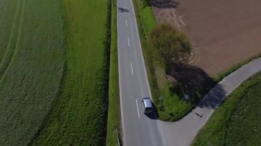 Arka planda çimen tarlası olan bir araba yolda ilerliyor. Yol ağaçlarla kaplı ve gökyüzü açık.