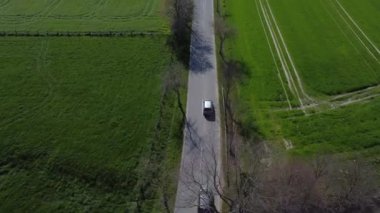 Kırsal kesimde bir araba yolda ilerliyor. Yol ağaçlar ve otlarla çevrili.