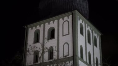 Yeşil çatılı ve beyaz duvarlı yüksek bir bina. Bina gece aydınlanıyor. Sahne gizemli ve ürkütücü.