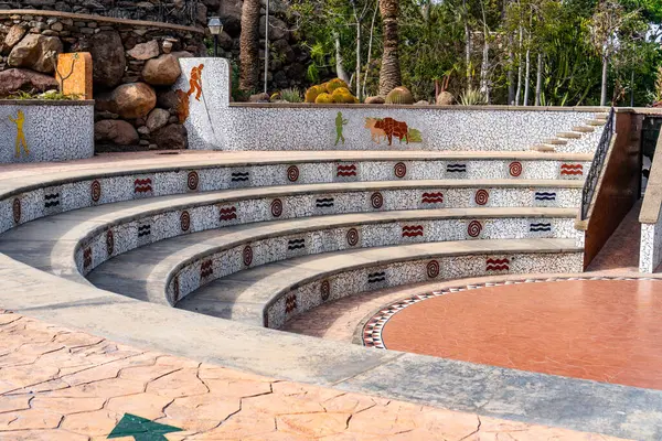 Mozaik tasarımlı ve ortasında taş bir çeşme olan bir taş geçit. Yürüme yolu kıvrımlı ve oraya giden basamakları var.