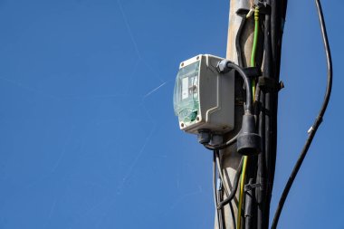 Bir elektrik kutusu, içinden kablolar çıkan bir direğin üzerinde. Gökyüzü mavi ve berrak