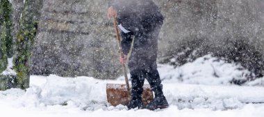 Bir adam kürekle kar kürüyor. Kar yağıyor ve adam siyah bir ceket giyiyor.