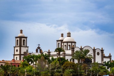 Bir sürü penceresi ve yüksek kulesi olan büyük beyaz bir bina. Bina ağaçlarla çevrili ve bir sürü yeşil yaprakları var. Gökyüzü mavi ve arka planda bazı bulutlar var