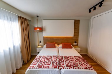 Beyaz yataklı, kırmızı ve turuncu yatak örtülü bir yatak odası. Yatak hazır, oda temiz ve düzenli.