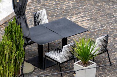 Önünde dört sandalye ve saksı bitkisi olan siyah beyaz bir masa. Masa tuğla bir duvar ve siyah bir şemsiye ile çevrilidir. Sahne sakin ve sakin.