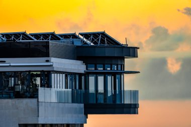 Şehre bakan camdan balkonlu bir bina. Balkon batan güneşle aydınlanır ve sıcak ve davetkar bir atmosfer yaratır.