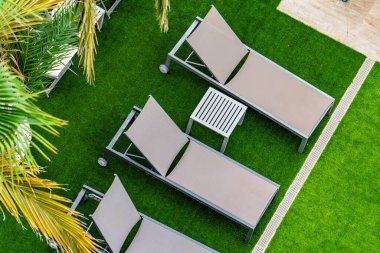 Bir sıra şezlong yemyeşil çimlerin üzerine kurulmuş. Sandalyeler sıraya dizildi, bir sandalye kameraya dönük ve diğerleri uzağa bakıyor. Sandalyelerin hepsi aynı renk.