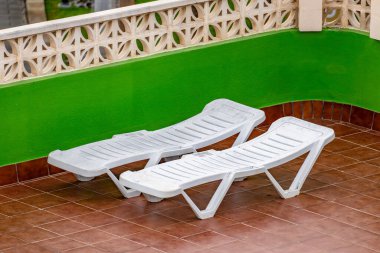 İki beyaz şezlong kahverengi fayans zemininde oturuyor. Sandalyeler birbirine bakıyor ve boş.