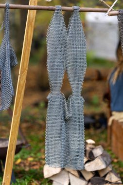 Ahşap bir direkten sarkan iki parça metal ağ. Ağ gümüşten ve pürüzlü bir dokusu var. Sahne açık havada, yakınlarda bir odun yığını var.