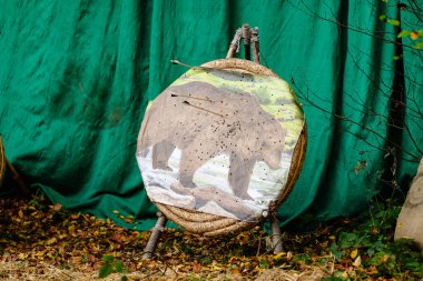 Yuvarlak bir hedef üzerinde ayı tasvir edilir ve oklar onu gösterir. Ayı yeşil bir perde ile çevrilidir ve yerde bazı yapraklar vardır. Avlanma ya da atış talimi kavramı