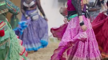 Bir grup kadın renkli kıyafetler içinde tarlada dans ediyor. Manzara canlı ve neşeli.