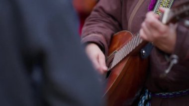 Bir kişi gitar çalıyor. Gitar kahverengidir ve siyah bir kafası vardır. Kahverengi bir ceket giyiyor.