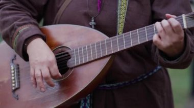 Bir kadın gitar çalıyor. Gitar eski moda bir enstrümandır. Kadın kahverengi bir ceket giyiyor.