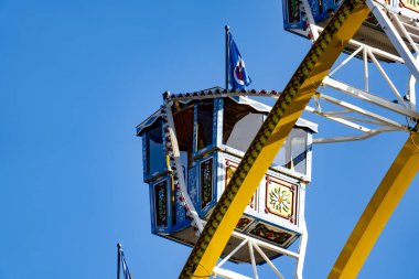 Üstünde küçük bir kulübe olan mavi ve sarı bir dönme dolap. Kulübe bayraklarla süslenmiş ve mavi bir çatısı var. Gökyüzü açık ve mavi