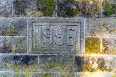 Üzerinde 1996 yazan taş plakalı tuğla bir duvar. Plaket yosunla kaplı ve yıpranmış görünüyor.
