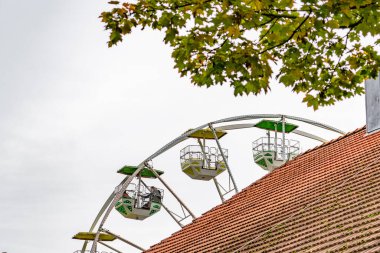 Kırmızı çatının üstünde üç arabalı bir dönme dolap var. Gökyüzü bulutlu ve ağaçtaki yapraklar yeşil