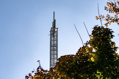 Arka planda önünde bir ağaç olan uzun bir kule görülür. Ağacın yaprakları renk değiştiriyor, resme sonbahar hissi veriyor. Gökyüzü açık ve mavi