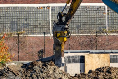 Sarı ve siyah bir inşaat makinesi toprağı kazıyor. Makine tuğla bir duvarla çevrili.