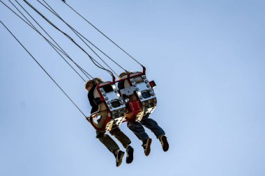 İki kişi salıncakta sallanıyor, biri siyah gömlek giyiyor. Gökyüzü açık ve mavi