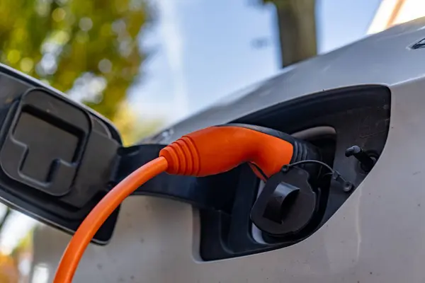 Bir araba elektrikle şarj ediliyor. Araba gümüş ve şarj kablosu turuncu. Araba bir otoparka park edilmiş.