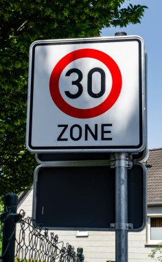 Üzerinde kırmızı daire ve beyaz numara 30 olan bir tabela. Tabela bir evin yanındaki direkte.