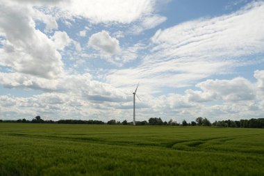 Bir rüzgar türbini çimen tarlasında duruyor. Gökyüzü bulutlu ve güneş bulutların arasında parlıyor.