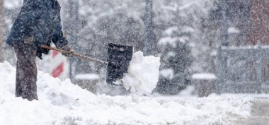 Bir adam kar küreme aracıyla kar kürüyor. Kar yağıyor ve adam mavi bir ceket giyiyor.