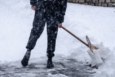 Bir adam tahta bir kürekle kar kürüyor. Kar yağıyor ve adam siyah pantolon giyiyor.
