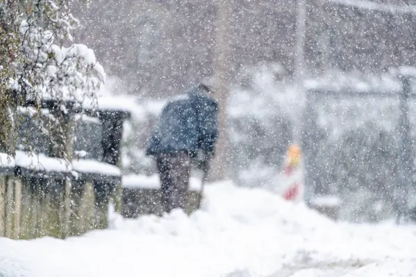 Bir adam kaldırımdan kar kürüyor. Kar yağıyor ve adam mavi bir ceket giyiyor.