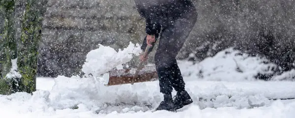 Bir adam kar fırçasıyla kar kürüyor. Kar yağıyor ve adam siyah bir ceket giyiyor.
