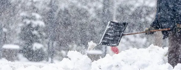 Bir adam kar küreme aracıyla kar kürüyor. Kar yağıyor ve gökyüzü bulutlu. Adam siyah bir ceket ve eldiven giyiyor.