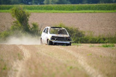 Bir araba toprak tarlasının içinden geçiyor. Araba bakımsız durumda, kaputu açık ve camları kırık. Olay yeri ıssız ve terk edilmiş. Etrafta başka araç ya da insan yok.