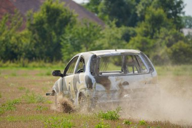 Bir araba toprak ve çimlerin arasından geçiyor. Araba çamurla kaplı ve ön tarafı hasarlı. Sahne kaotik ve dağınık. Araba arkasında toprak bırakıyor.