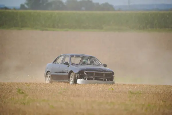 Siyah bir araba kuru otların arasından geçiyor. Araba hasar görmüş ve ön camı kırılmış.