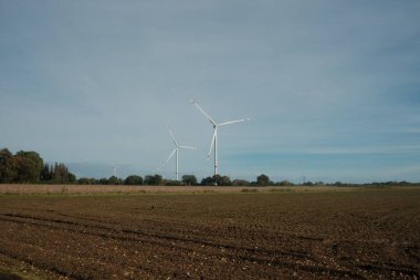 Arka planda iki rüzgar türbini olan bir alan. Gökyüzü açık ve güneş parlıyor.