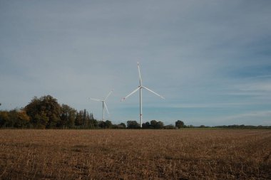 Uçsuz bucaksız, açık bir alanda iki rüzgar türbini dik duruyor. Gökyüzü bulutlu, sakin ve sakin bir atmosfer yaratıyor. Doğa ve teknoloji arasındaki uyum kavramı