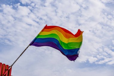Gökyüzünde bir gökkuşağı bayrağı dalgalanıyor. Bayrak renkli ve canlı, LGBTQ + toplumunu temsil ediyor. Gökyüzü açık ve mavi, birkaç bulut etrafa saçılmış.